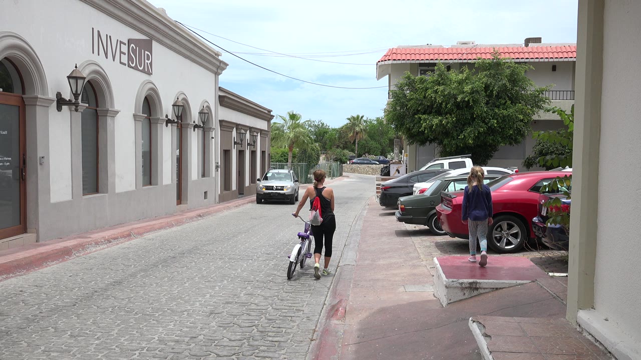 Woman and child with a bicycle on a quiet street beside parked cars.