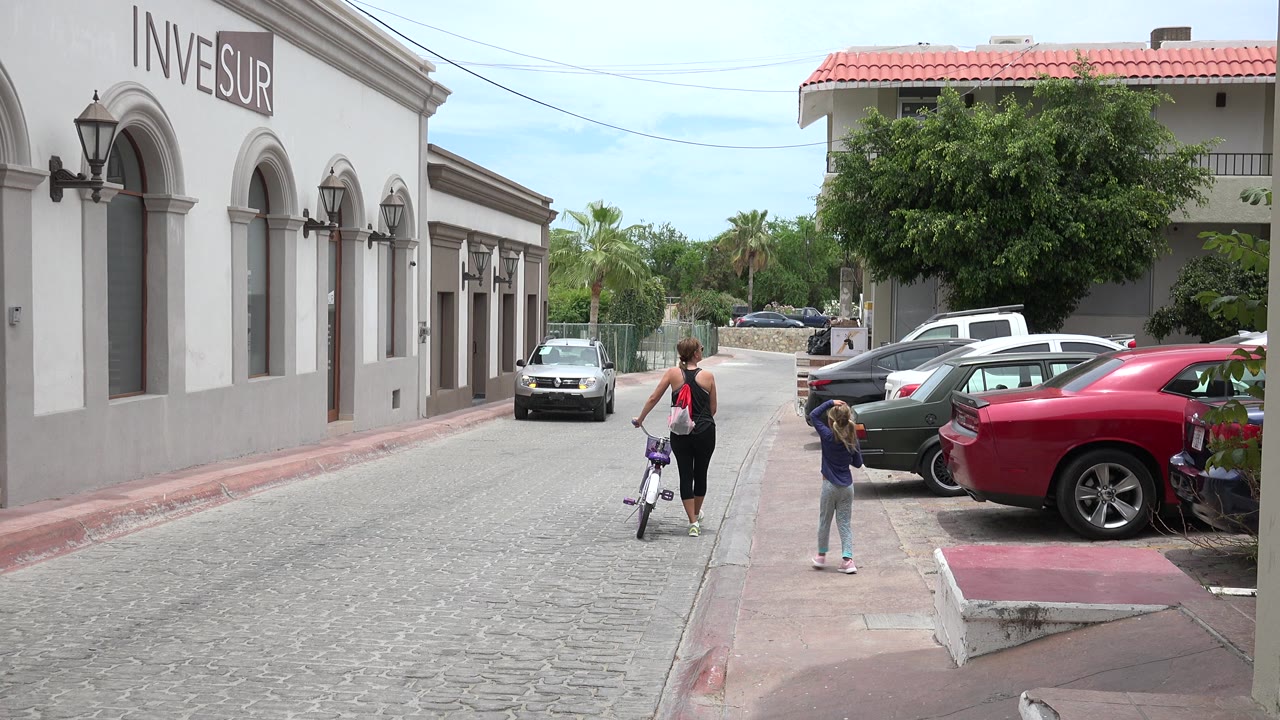 In Los Cabos, Mexico, a woman and young girl move away from the camera with a small bicycle on a paved street lined by arched buildings and parked cars.