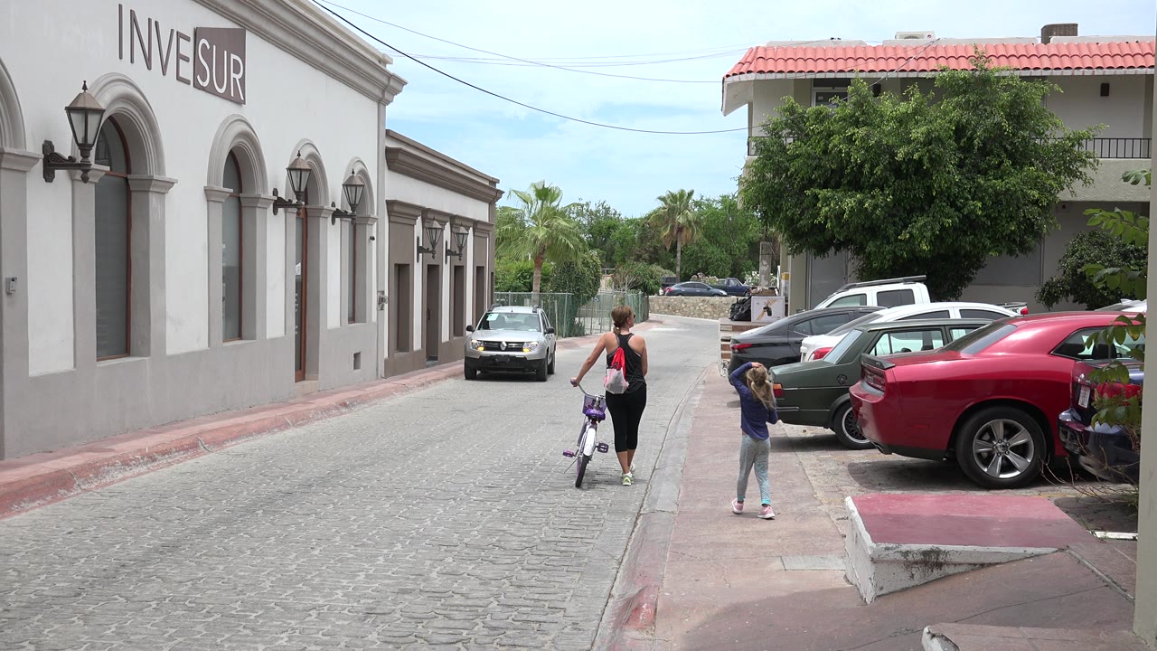 The woman and young girl are farther down the Los Cabos street, with the bicycle between them and a row of parked cars along the right side.