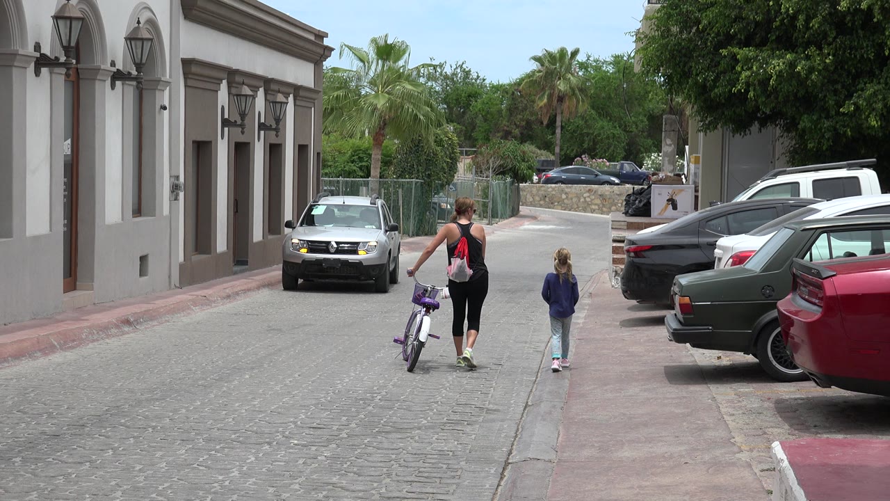 Woman walking a bicycle beside a child on a cobblestone street with cars parked nearby.