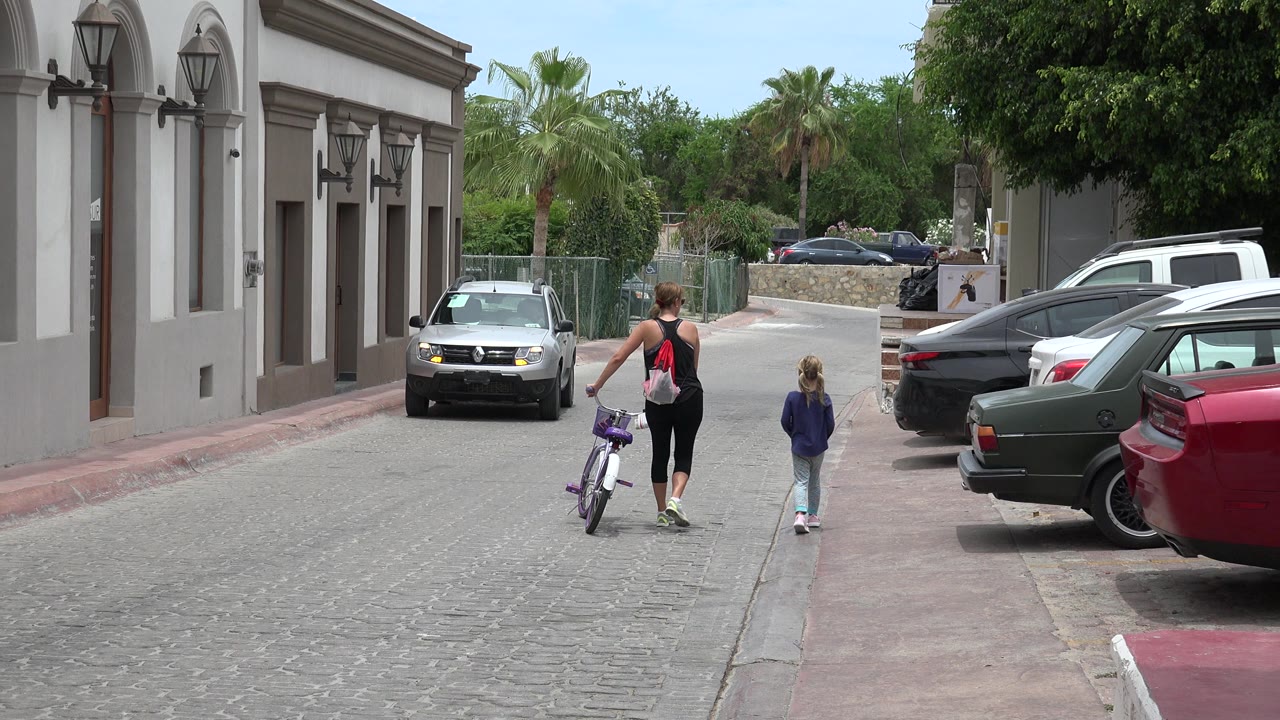 One second into the Los Cabos street scene, the woman with the bicycle and the child continue away from the camera as a car faces them from the lane ahead.