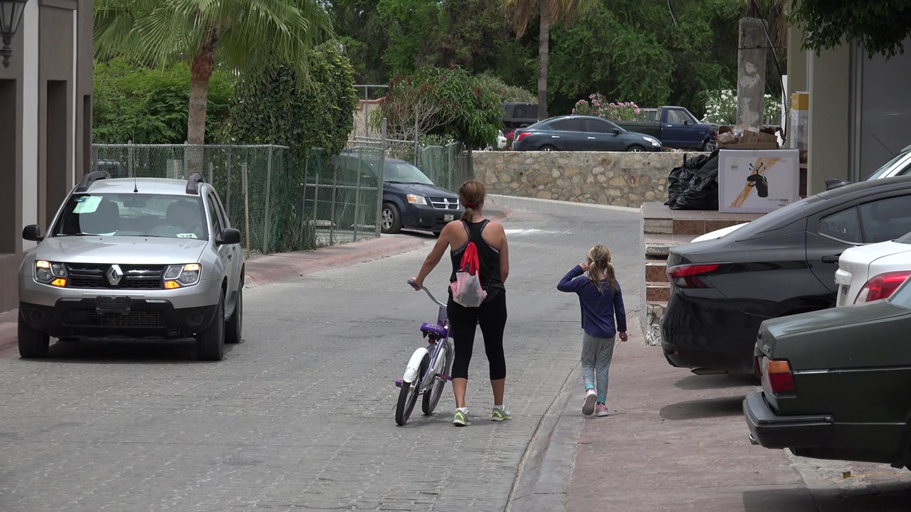 The woman and young girl move farther down the Los Cabos street, with a gray vehicle facing the camera and parked cars framing the road.