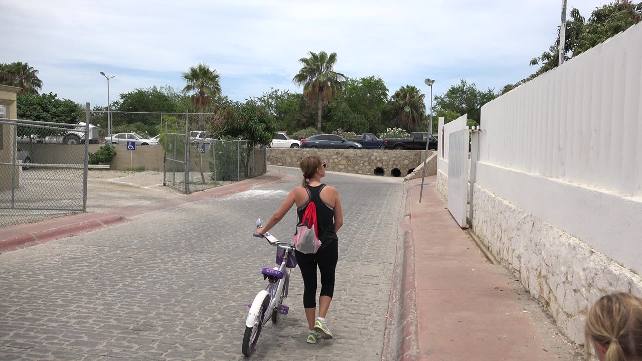 Woman pushing a bicycle down a paved street with a fence and white wall.