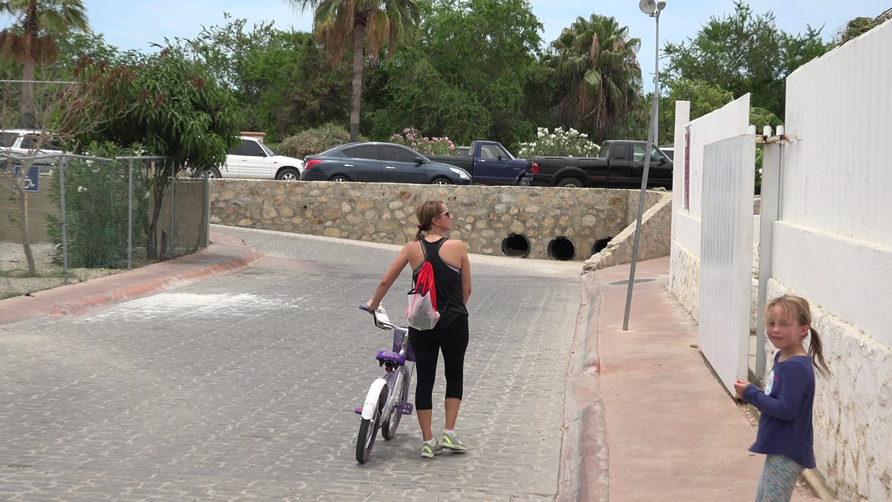 Woman walking a bicycle toward a bridge with a child near a white wall.