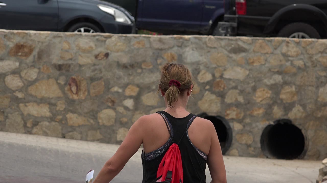 Woman seen from behind near a stone wall and parked vehicles.