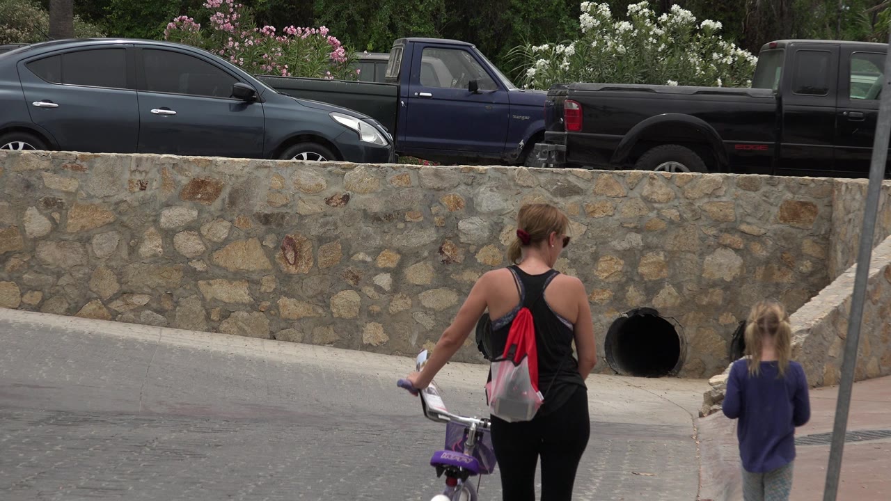 The woman and child move farther along the Los Cabos sidewalk, with the bicycle wheel near the curb and dark vehicles parked beyond the stone wall.