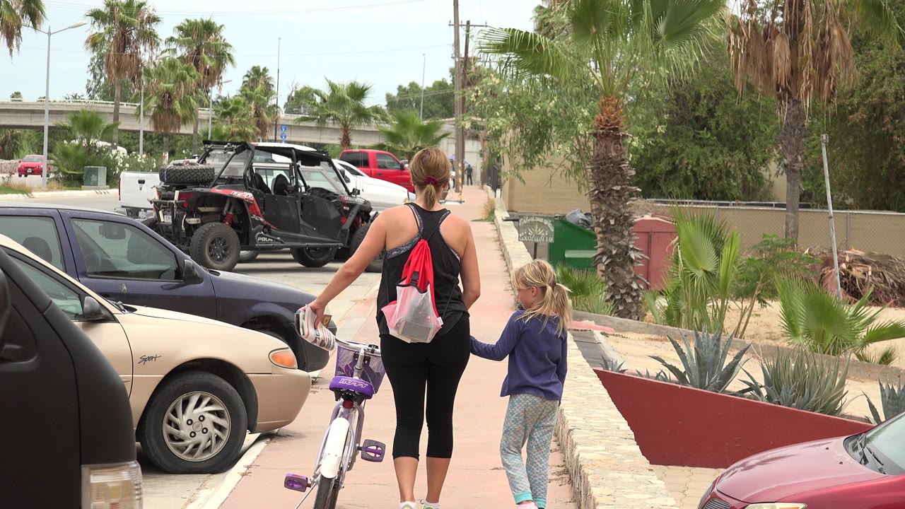 In Los Cabos, Mexico, a woman and young girl walk away from the camera on a sidewalk, with a small purple bicycle beside them and parked cars along the curb.