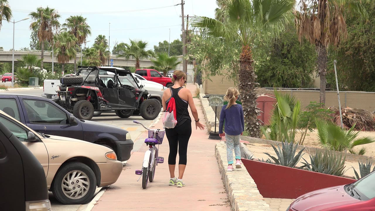 At the start of the Los Cabos clip, a woman walks away from camera beside a purple bicycle while a young girl walks ahead near parked cars, palm trees, and desert landscaping.