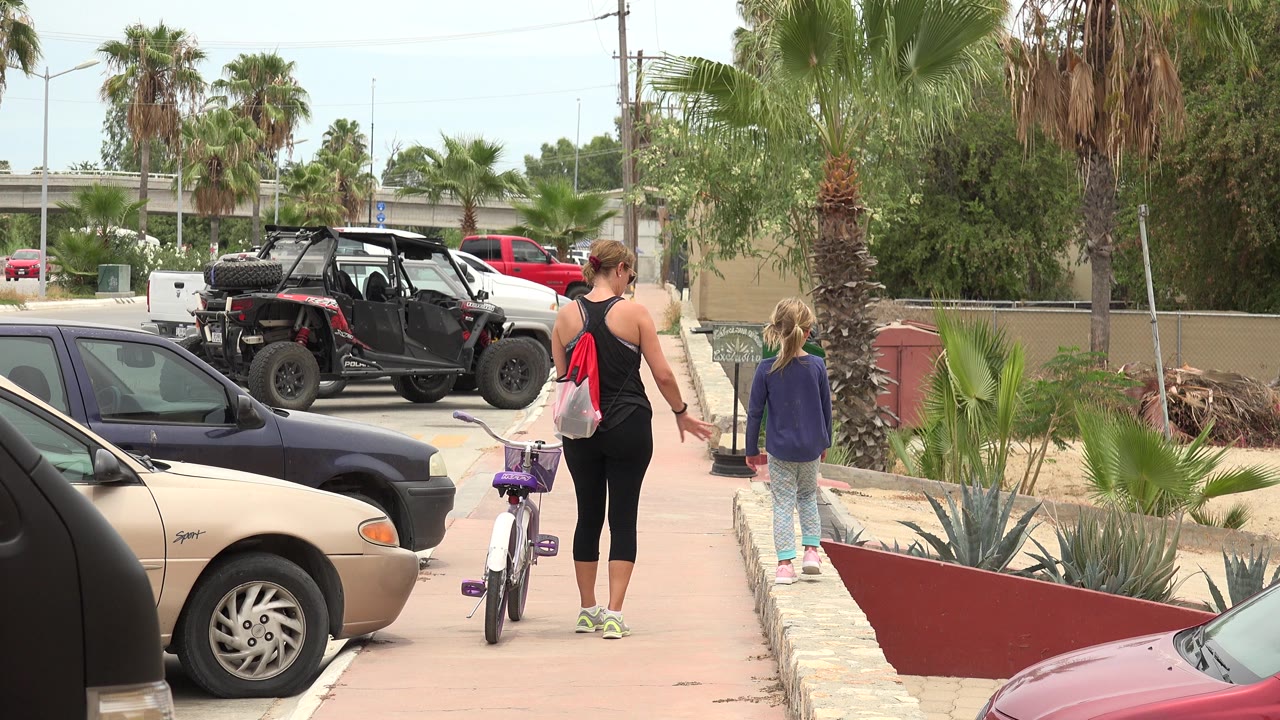 One second in, the woman and child continue away from camera along the Los Cabos sidewalk, with the bicycle closer to the center and parked vehicles lining the street edge.