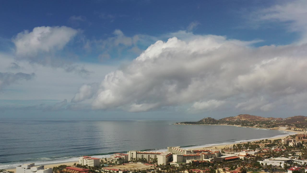 Opening frame of Los Cabos, Mexico, showing calm ocean at left, a curving beach and low shoreline buildings below, distant hills at right, and no visible people.