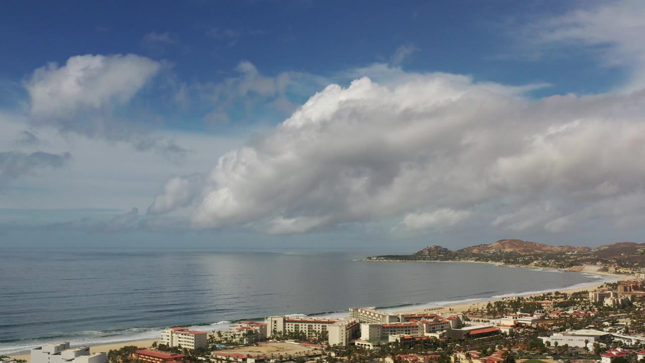 Elevated beach and ocean view with hills, buildings, and broad cloud cover.