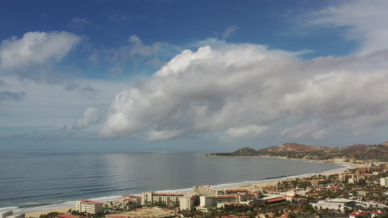 Wide elevated view of Los Cabos, Mexico with a blue ocean bay, curving beach, coastal buildings, brown hills, large white clouds, and no visible people.