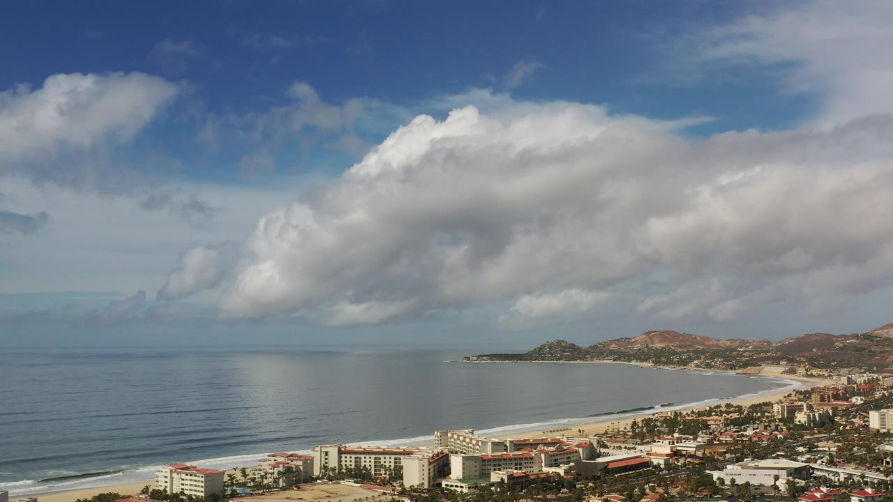Blue ocean, beach, buildings, hills, and clouds.