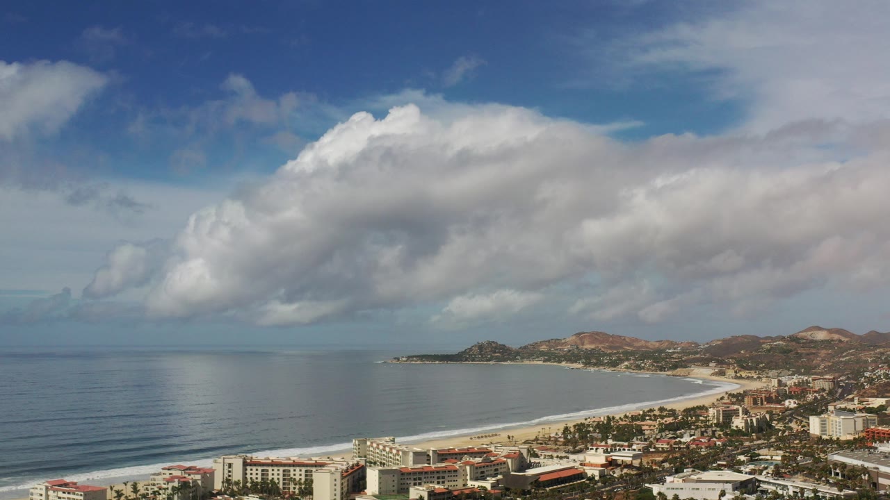 Elevated coastline view with ocean, beach, buildings, hills, and clouds.