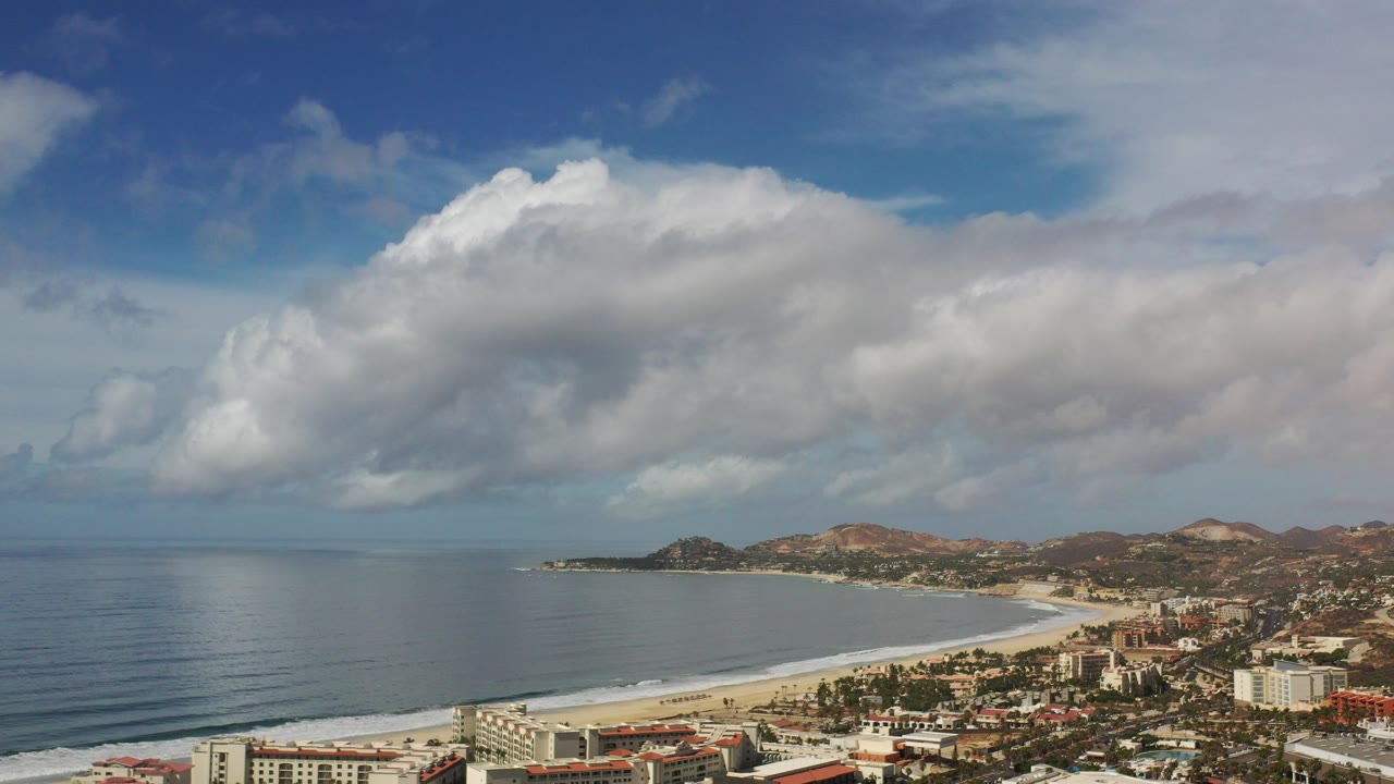 Wide aerial view of ocean, beach, coastal buildings, hills, and large clouds.