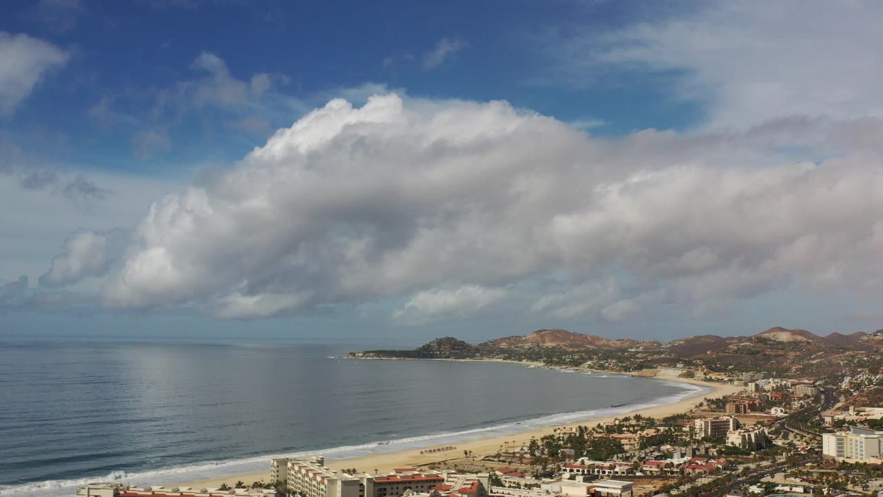 Opening frame of the Los Cabos coast from an elevated view, with dark ocean at left, a curved pale beach, low buildings at lower right, hills beyond, and no visible people.