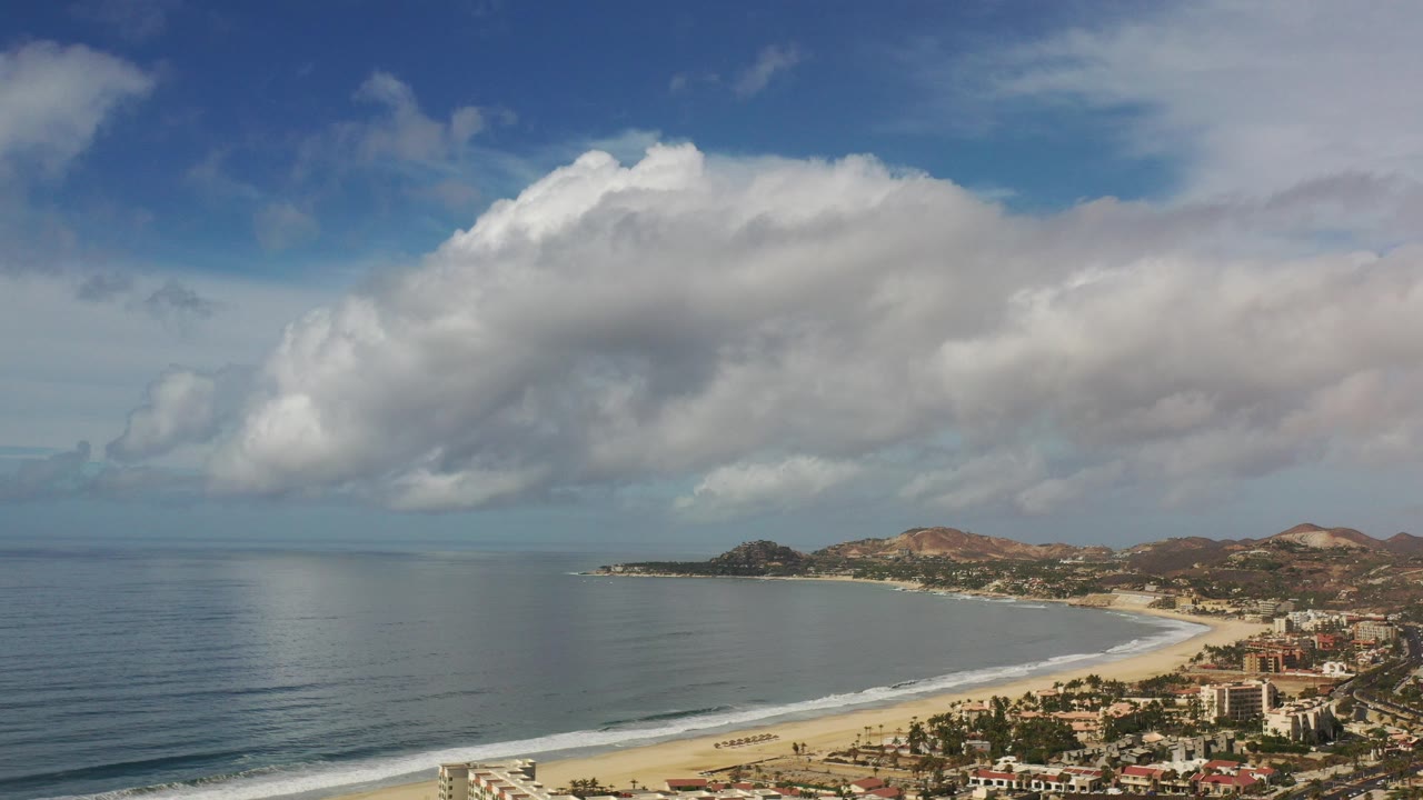 Aerial view in Los Cabos, Mexico, with calm ocean at left, a curved sandy shoreline at right, coastal buildings below the hills, broad cloud cover, and no visible people.