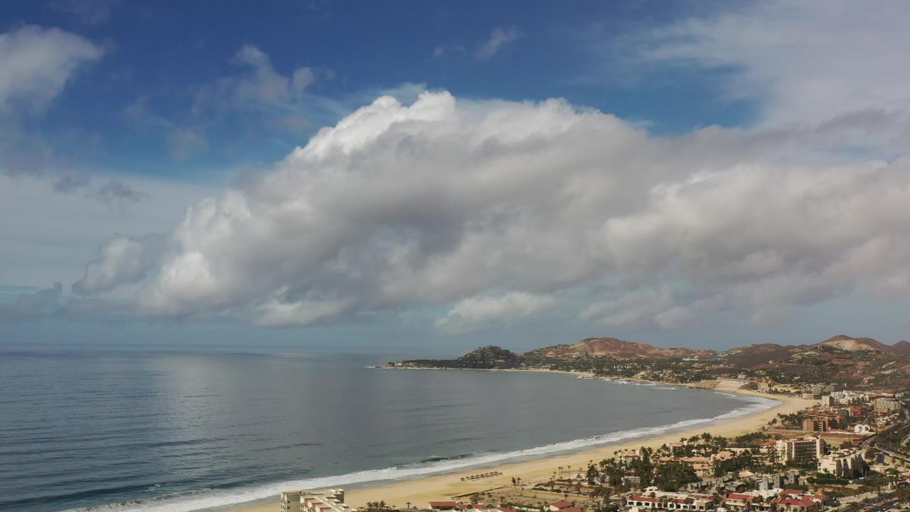 Wide coastal aerial with ocean, beach edge, buildings, hills, and bright clouds.