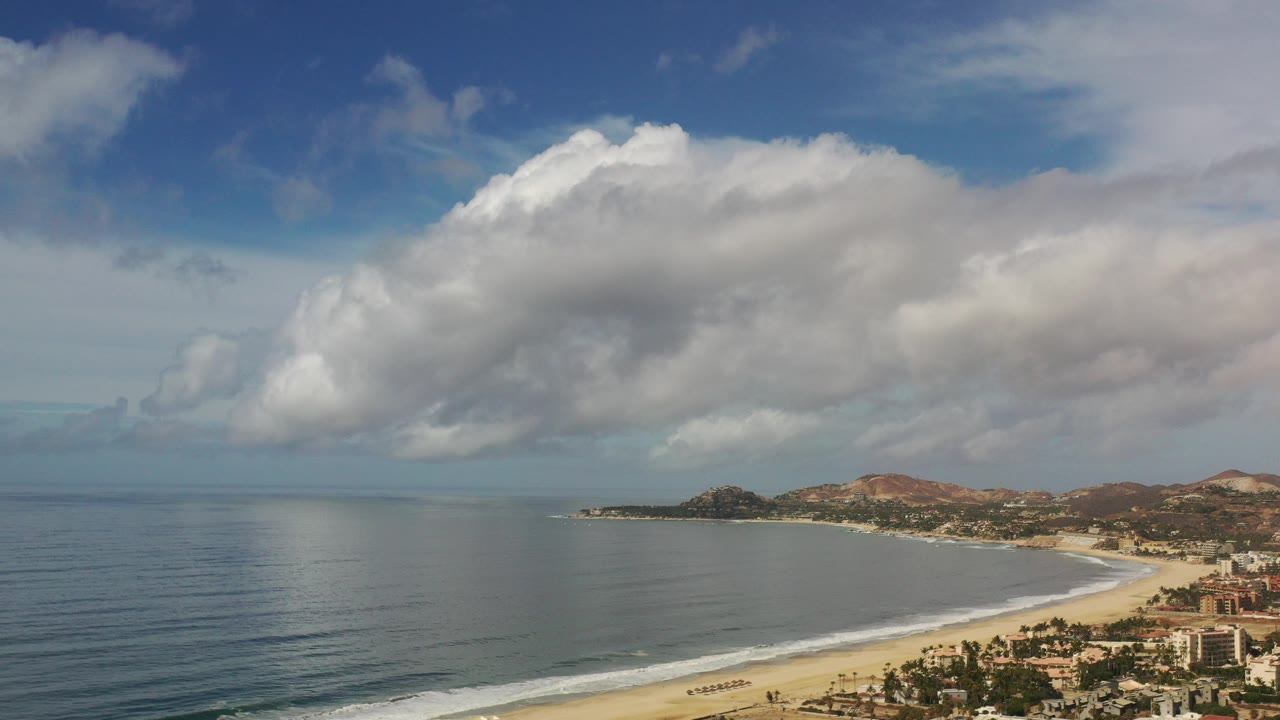 Wide aerial view of a curved beach, ocean, clouds, hills, and coastal buildings.