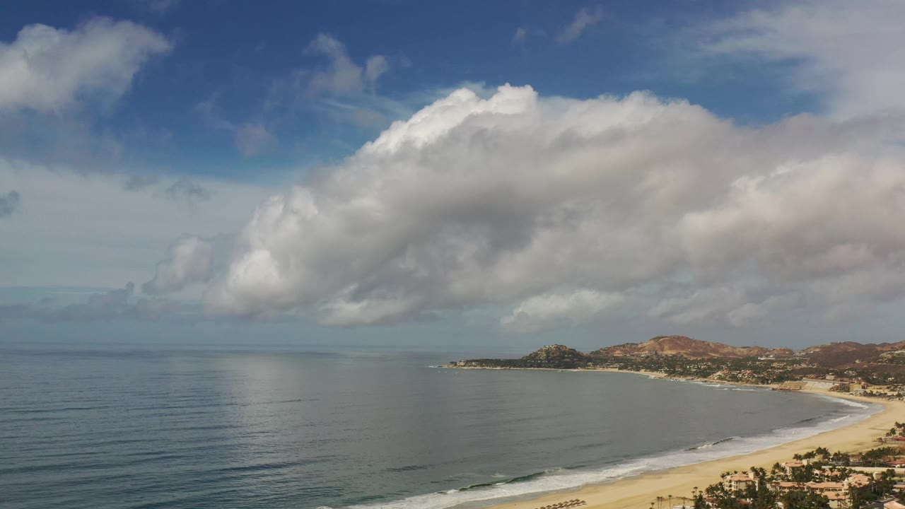 Los Cabos shoreline seen from above with broad ocean water, a pale beach along the right edge, mountain ridges beyond, thick white clouds, and no visible people.