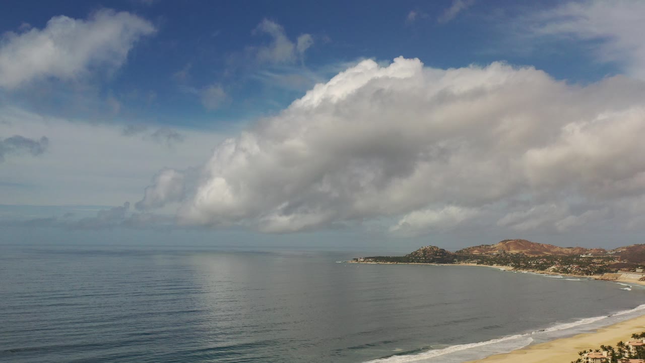 Wide ocean, sandy shoreline, rocky point, hills, and large clouds.