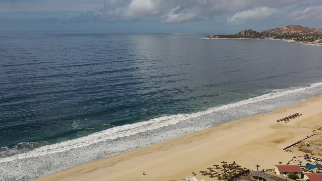 Aerial view in Los Cabos, Mexico with rippled blue water taking most of the frame, a diagonal surf band, tan beach, grouped umbrellas, and small beachfront structures below.