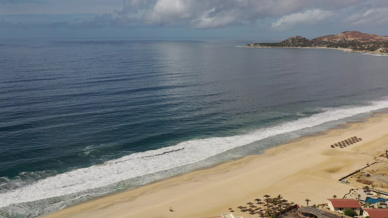 Opening Los Cabos, Mexico aerial frame with dark blue ocean on the left, white surf crossing the lower shoreline, beach umbrellas near the sand, distant coastal hills, and no clearly identifiable people.