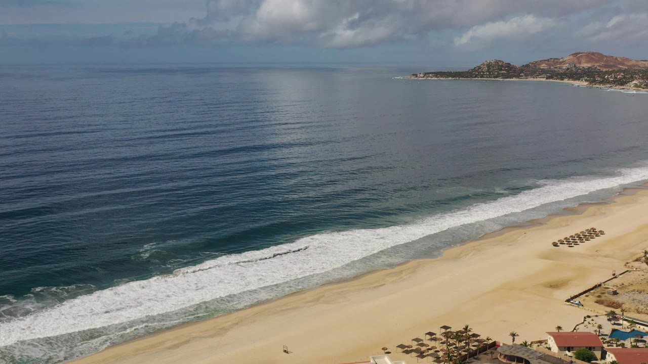 Los Cabos beach aerial at 0:01 with a broad expanse of ocean, a diagonal white wave band, pale sand, small umbrella rows, cloudy blue sky, and no clearly identifiable people.