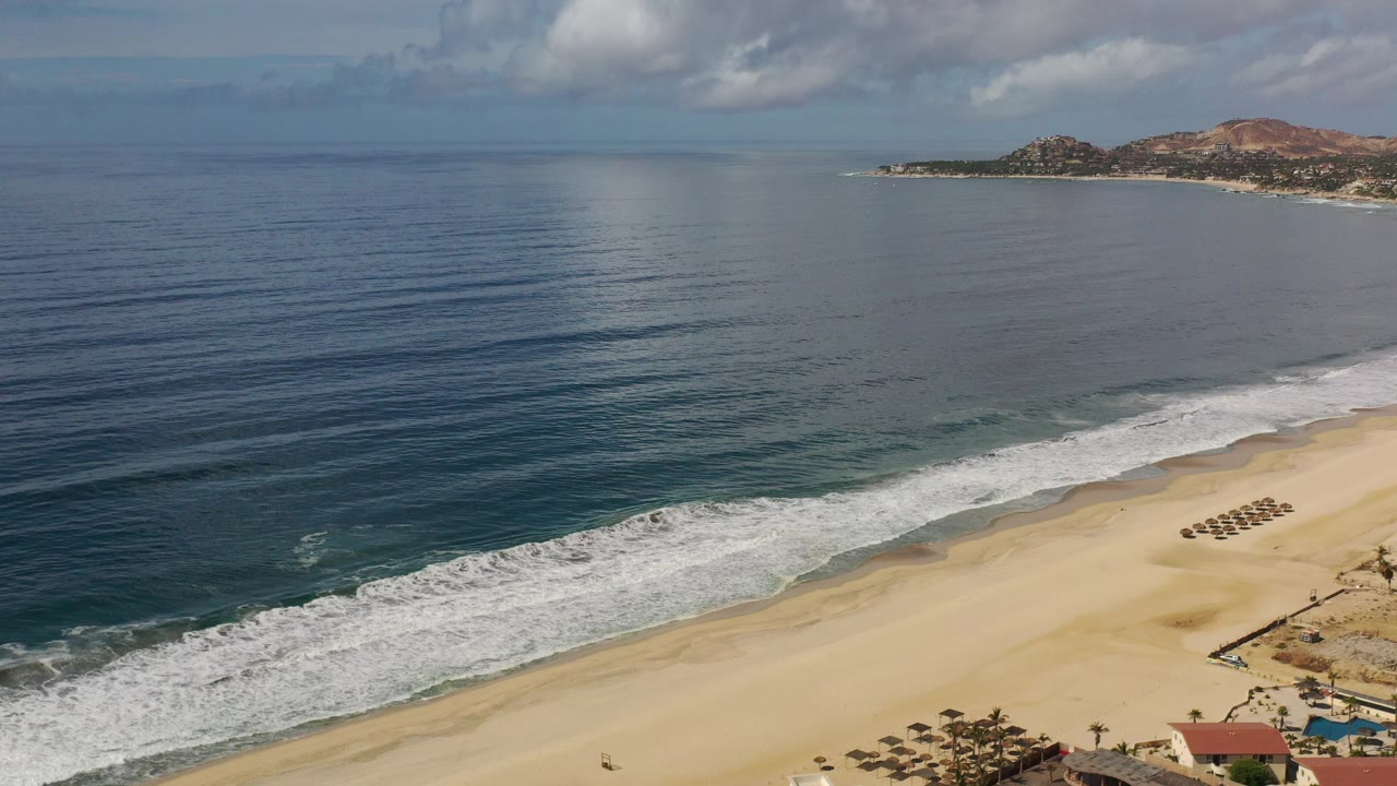 Aerial view of blue ocean, surf, sandy beach, and distant rocky coastline.