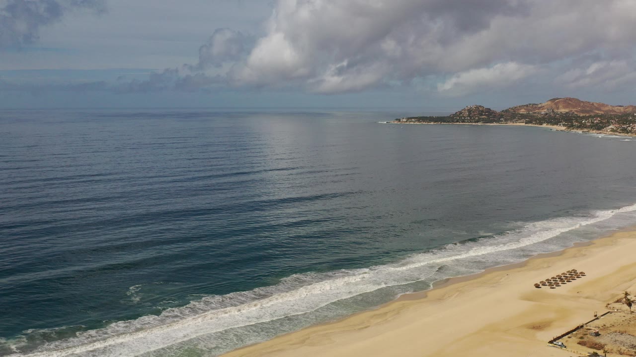 Elevated view of the Los Cabos coastline with dark blue ocean, white surf, sandy beach at lower right, distant brown hills, gray cloud cover, and no visible people.