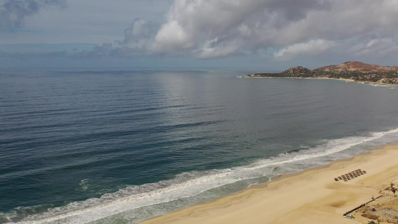 Wide Los Cabos coastal frame with parallel wave lines moving toward the sand, a small cluster of beach umbrellas or loungers, distant hills on the right, and no visible people.