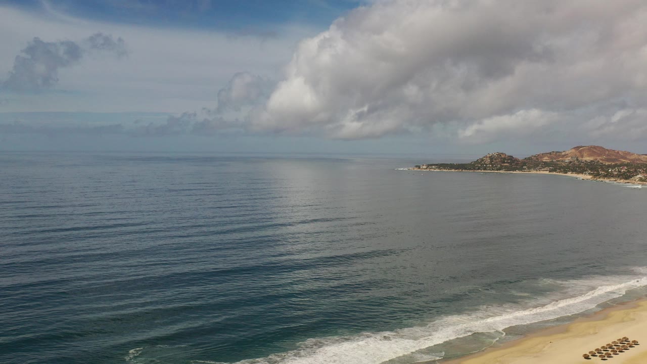 Wide view over the Los Cabos ocean with rippled blue water, large gray-white clouds, distant rocky coastline at right, beach surf at lower right, and no visible people.