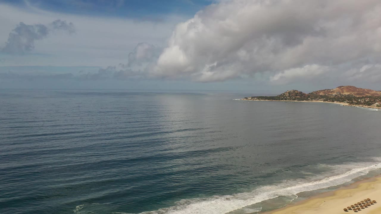 Los Cabos coastal frame with calm water bands, a bright patch of reflected light, broad cloud cover overhead, sandy beach at the lower right, and no visible people.