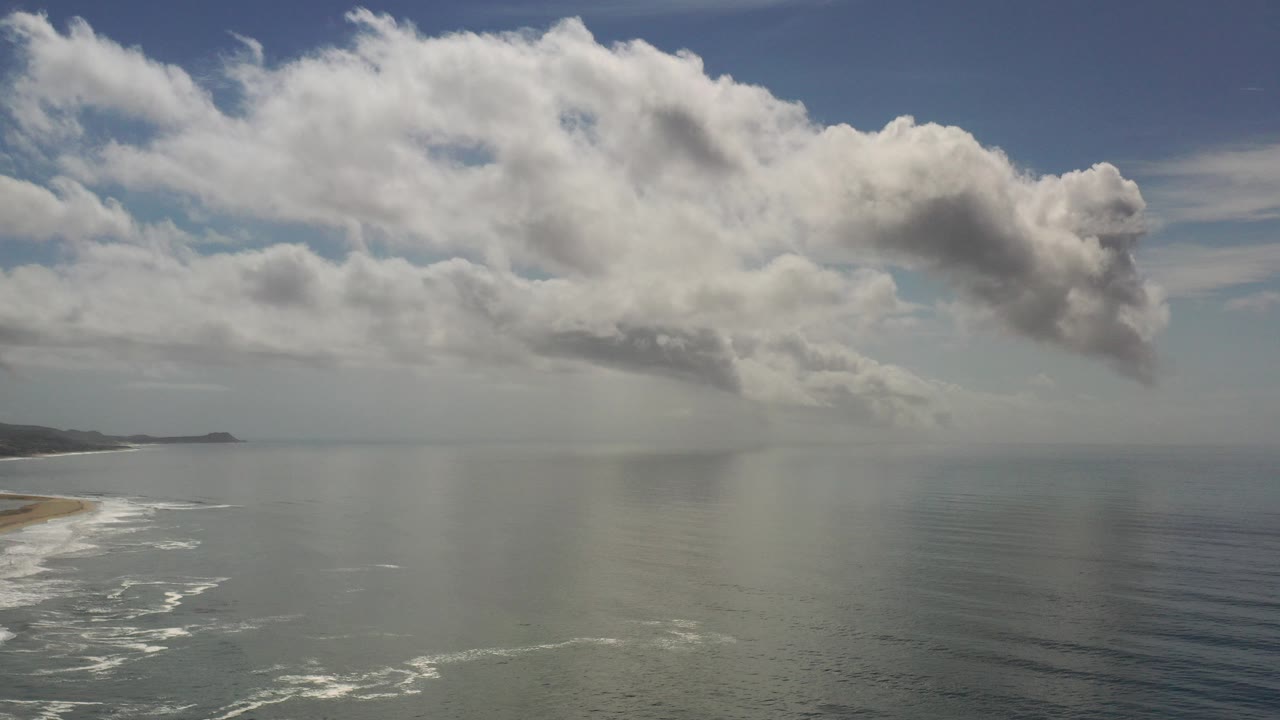 Opening frame of a Los Cabos ocean aerial with sandy shoreline at left, broad water below, large cumulus clouds overhead, distant land on the horizon, and no visible people.