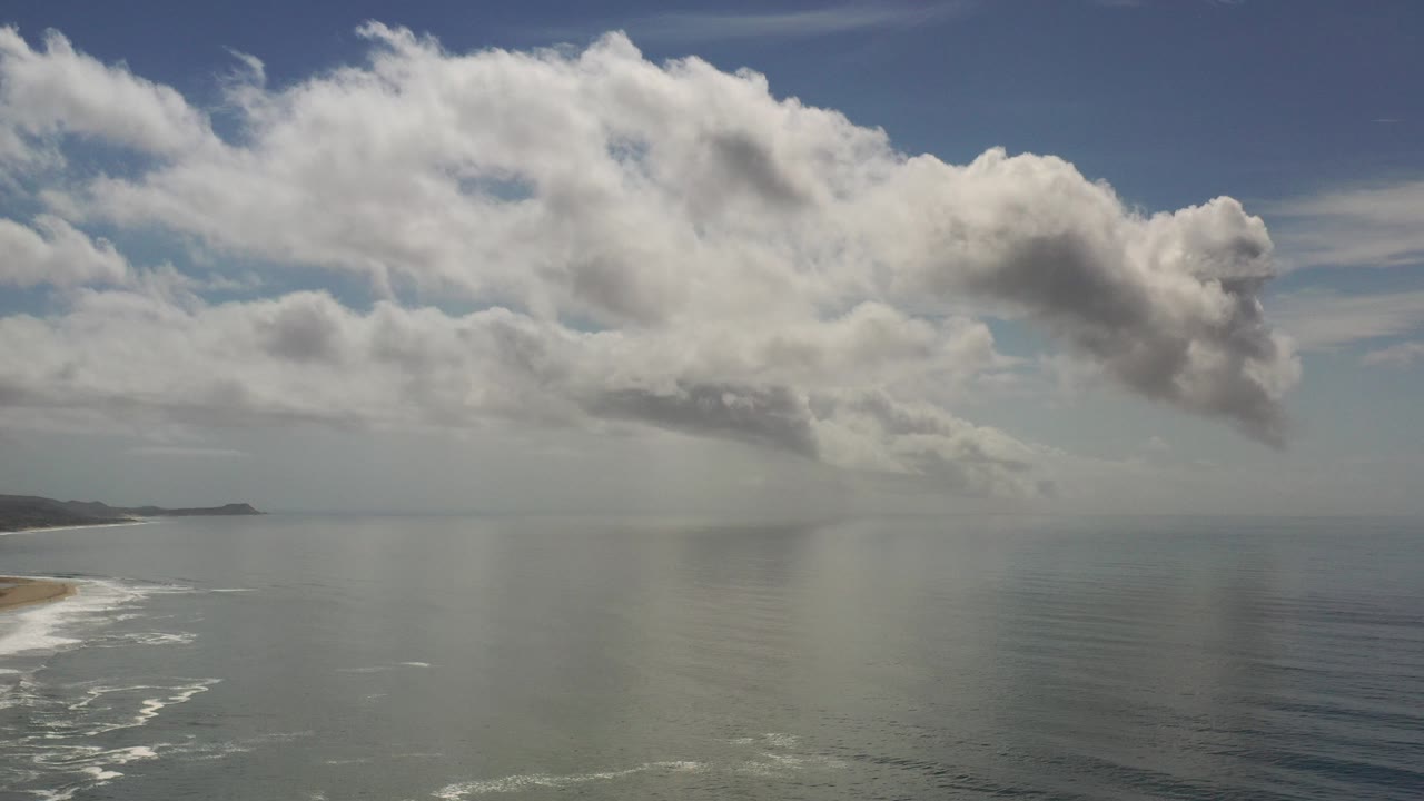 Opening frame of Los Cabos ocean under broad low clouds, with pale surf along the lower-left beach and no visible people.
