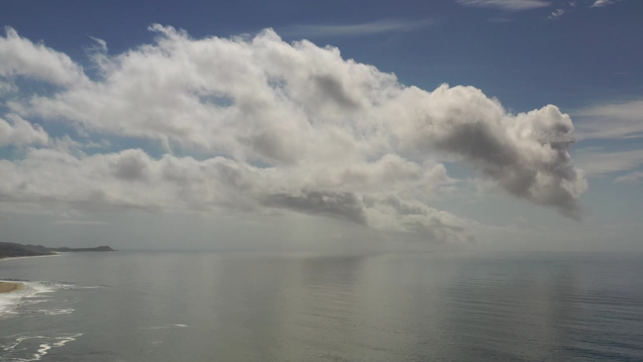 Los Cabos, Mexico aerial view over open ocean with broad white clouds, faint rain haze over the water, surf along the left shoreline, and no visible people.