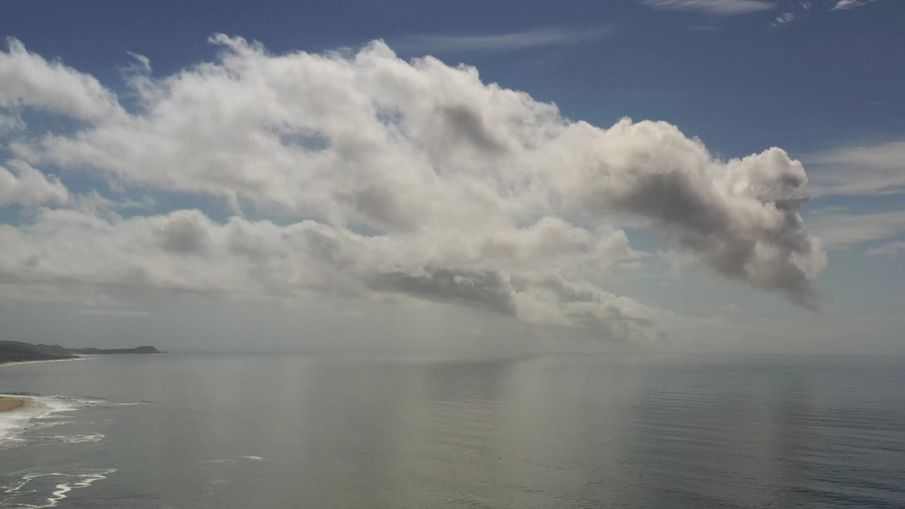 Los Cabos, Mexico coastline remains at the lower left while layered clouds stretch across the sky above calm ocean water, with no visible people.