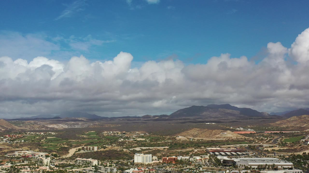 Wide aerial view of city buildings, desert land, mountains, and clouds.