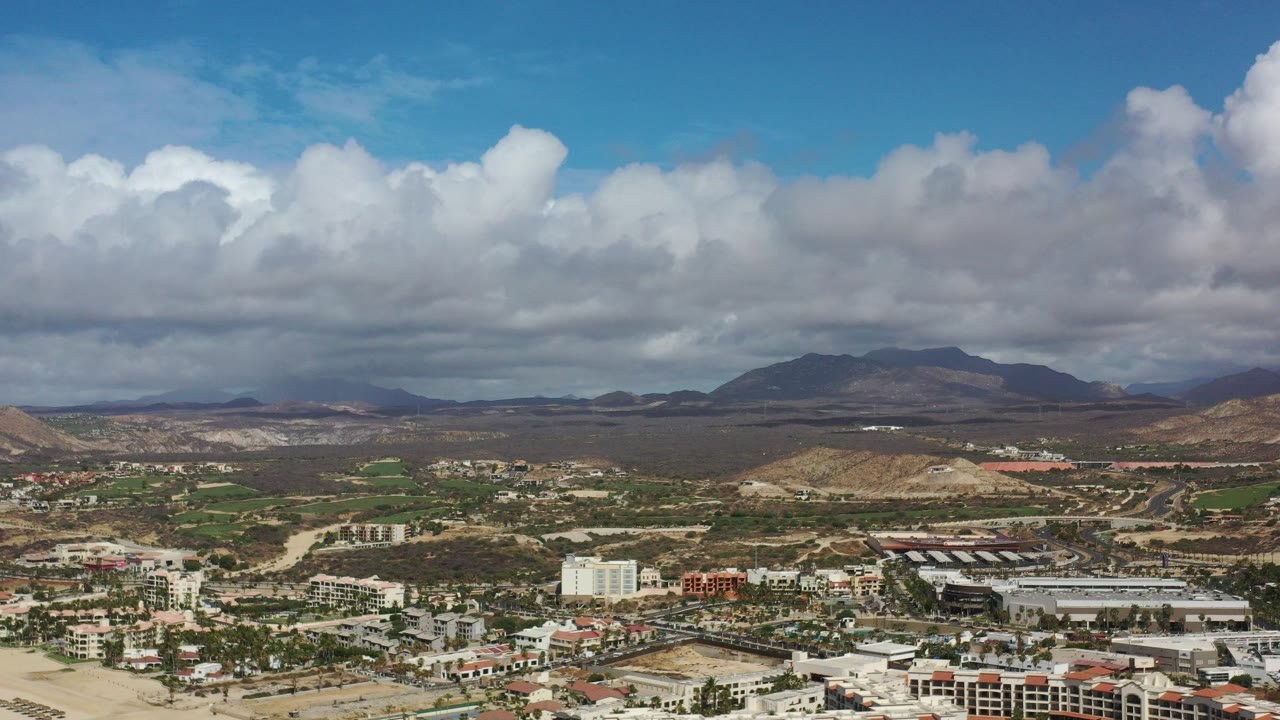 Opening aerial frame over Los Cabos, Mexico, showing low-rise town buildings, roads, dry hills, distant mountains, thick clouds, and no visible people.