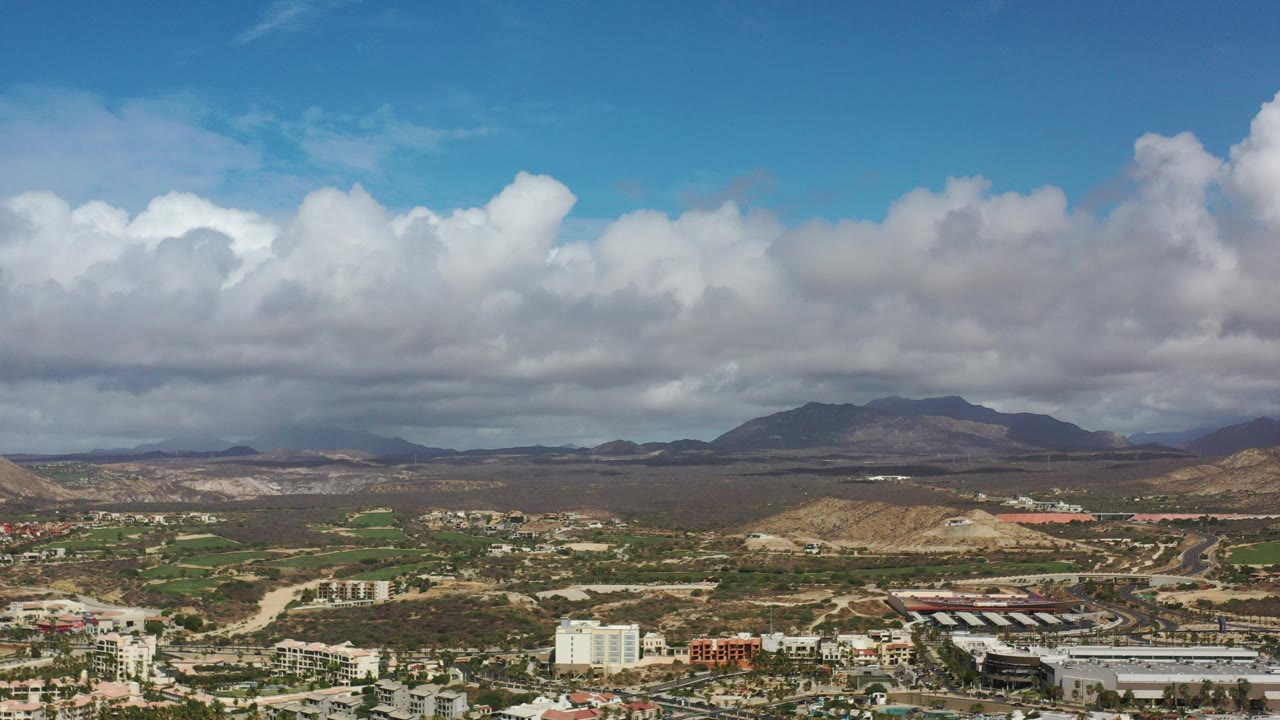 Opening view over Los Cabos, Mexico, with white clouds above distant mountains, dry terrain, green patches, roads, and no visible people.