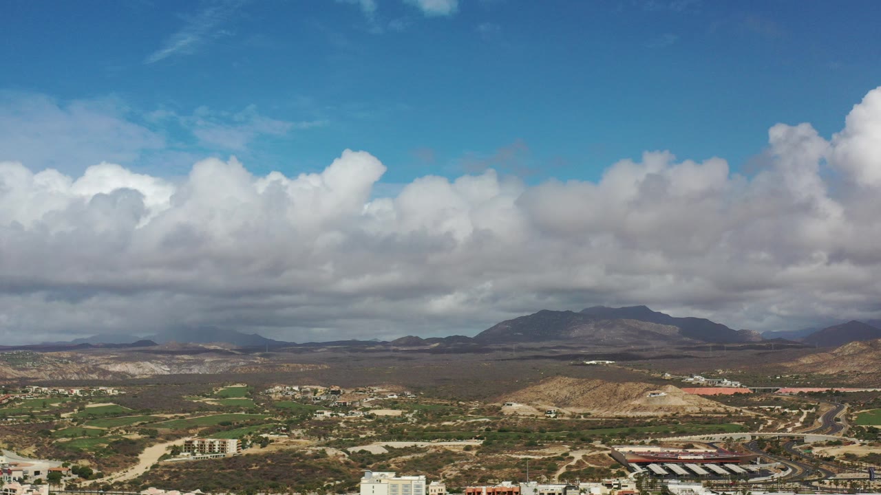 Wide aerial view of clouds over mountains, desert terrain, green areas, roads, and buildings.