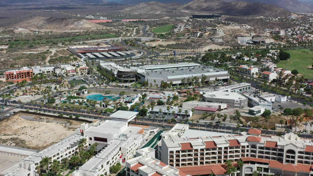 High aerial frame over Los Cabos, Mexico showing white resort-style buildings in the foreground, commercial structures and green fields beyond, brown mountains in back, and no visible people.