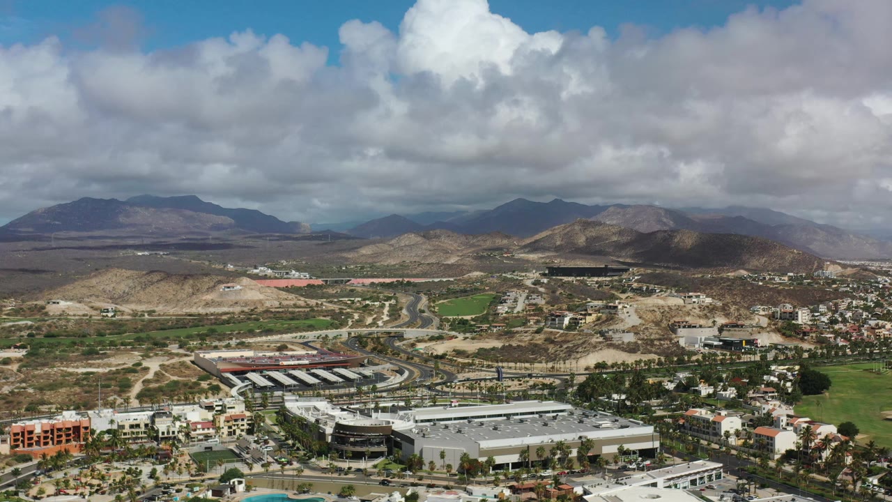 Early aerial view in Los Cabos, Mexico, showing a large light-roofed building, road network, scattered homes, dry hills, mountains, and no visible people.
