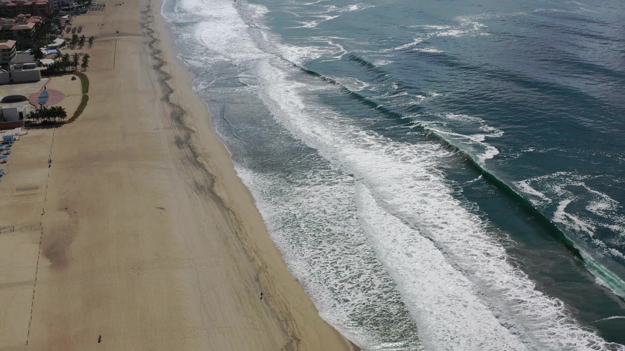A Los Cabos beach seen from an elevated angle, with a long white wave band pushing closer to the sand and darker blue water beyond, with no visible people.