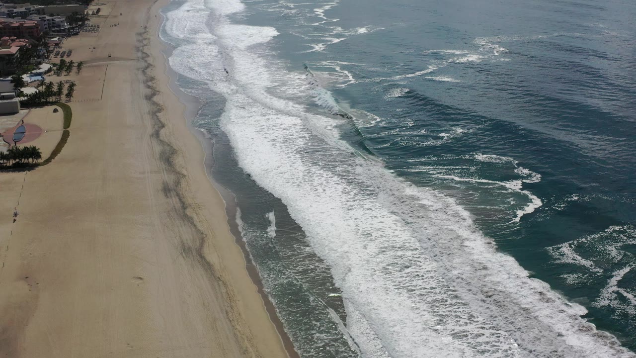 Aerial frame of the Los Cabos shoreline where white waves run diagonally across the center, broad sand stays clear on the left, and open ocean fills the right with no visible people.