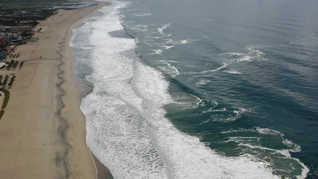 Opening aerial frame over Los Cabos, Mexico, showing white surf bands rolling beside a wide empty sand beach with no visible people.