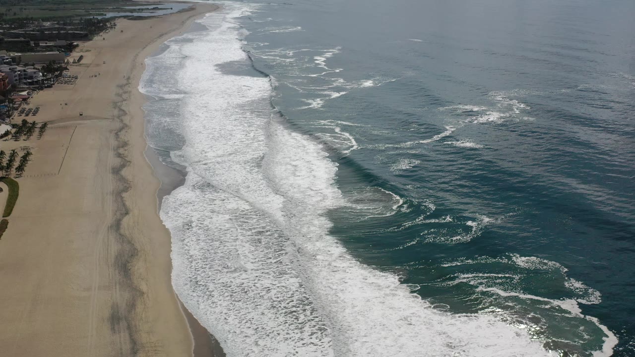 Aerial frame from Los Cabos, Mexico, with a broad foam field pushing onto the sand and open teal water to the right, with no visible people.