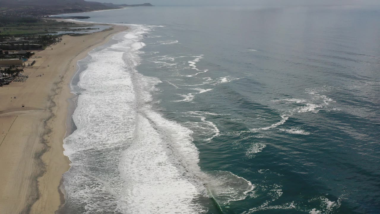 Opening aerial frame over a Los Cabos beach, with pale sand along the left edge, white surf lines down the center, blue-green ocean to the right, distant hills, and no visible people.