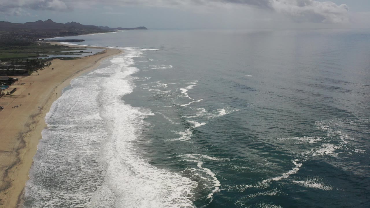 High aerial view over a Los Cabos, Mexico coastline with sandy beach on the left, white surf across the center, open ocean to the right, distant mountains, and no visible people.