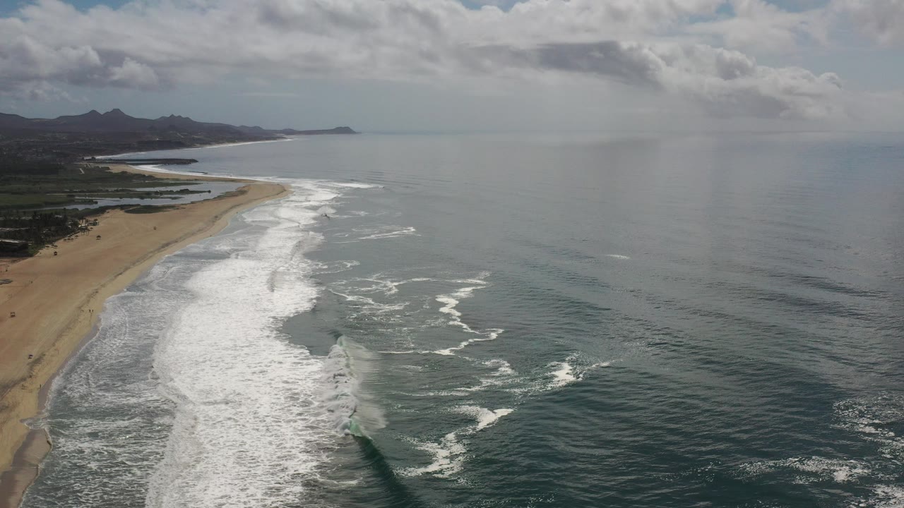 Aerial view of surf breaking along a sandy beach with ocean to the right.